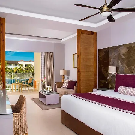 Dreams Royal Beach Punta Cana (Adults Only) Room photo The photo shows a bright and airy hotel room with a modern and luxurious feel. The main focus is a large bed with a white duvet, accented by a rich burgundy runner and several plush pillows in varying shades of purple and white with intricate pattern