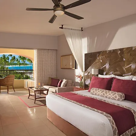 Dreams Royal Beach Punta Cana (Adults Only) Room photo The photo shows a beautifully decorated hotel room with a focus on a large bed and a balcony overlooking a tropical scene. 

The room features a king-sized bed with crisp white linens, several pillows including deep red and patterned decorative pillo