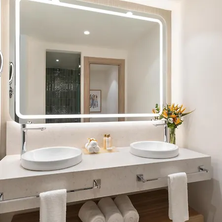 Dreams Royal Beach Punta Cana (Adults Only) Sinks photo The photo shows a modern and well-lit bathroom vanity area. The focal point is a large, rectangular mirror with a soft, illuminated border. The mirror reflects a glimpse into another part of the bathroom, showing a shower area with dark, textured wal