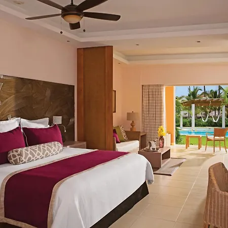 Dreams Royal Beach Punta Cana (Adults Only) Room photo This is a bright and airy photograph of a luxurious hotel bedroom with a stunning view of a tropical pool area. 

The focal point is a large king-sized bed with a dark wood headboard featuring an abstract, textured design. The bed is dressed in crisp