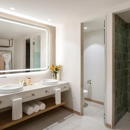 Dreams Royal Beach Punta Cana (Adults Only) Business photo The photo shows a bright and modern bathroom with a clean, minimalist design. The focal point is a large, rectangular mirror with a soft, illuminated frame above a long, white countertop. The countertop holds two sleek, white vessel sinks and a vase 