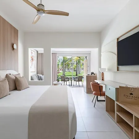 Dreams Royal Beach Punta Cana (Adults Only) Room photo The photo shows a bright and airy hotel room with a modern design. The focal point is a large bed with white linens and several light brown pillows. A light brown runner is placed across the foot of the bed. 

The room features a wooden headboard wit