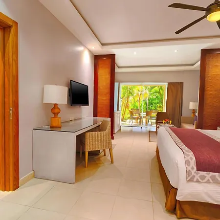 Dreams Royal Beach Punta Cana (Adults Only) Room photo The photo shows a bright and airy hotel room with a contemporary tropical vibe. The room is spacious and well-lit, with light-colored tiled floors. 

The main focus is a large bed with white linens and a rich maroon bed runner. Decorative pillows in 